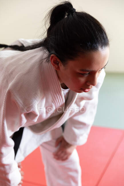 Vista lateral de cerca de una adolescente jugadora de judo femenino de raza mixta enfocada que usa judogi blanco, respirando y descansando durante un combate en un gimnasio. - foto de stock