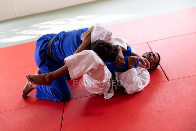 Vista trasera de alto ángulo de un entrenador de judo masculino de raza mixta y judoka masculino de raza mixta adolescente con judoka azul y blanco, practicando judo durante un entrenamiento en un gimnasio. - foto de stock