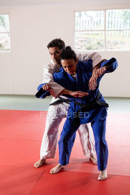 Vue de face d'un entraîneur de judo mixte masculin et d'une judoka mixte adolescente, portant du judogi bleu et blanc, pratiquant le judo lors d'un entraînement dans une salle de gym. — Photo de stock