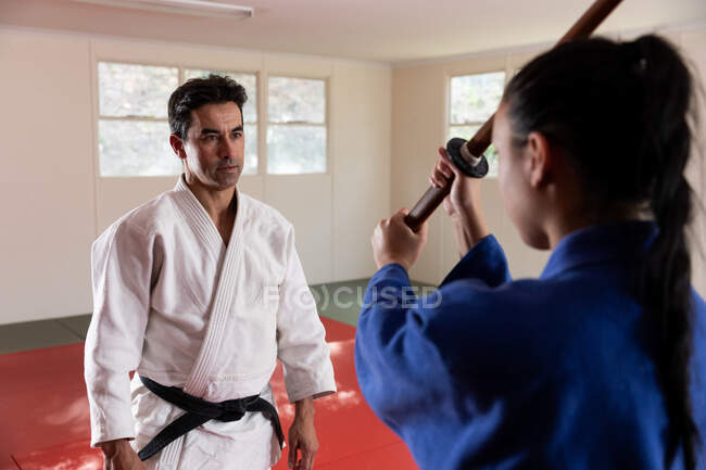 Vista trasera de una judoka femenina adolescente de raza mixta que usa judogi azul, practicando con un palo de judo jo durante un entrenamiento con un entrenador en un gimnasio. - foto de stock