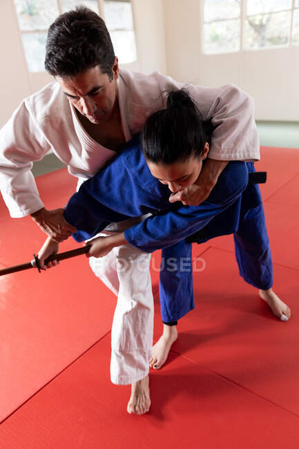 Vista frontal de cerca de una judoka femenina adolescente de raza mixta que usa judogi azul, practicando con un palo de judo jo durante un entrenamiento con un entrenador masculino en un gimnasio. - foto de stock