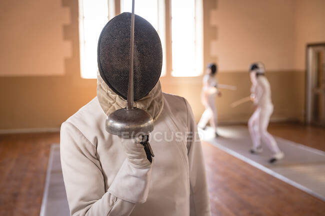 African American sportsman wearing protective fencing outfit during a fencing training session, preparing for a duel, holding an epee in front of face. Other fencers training in the background. — Stock Photo