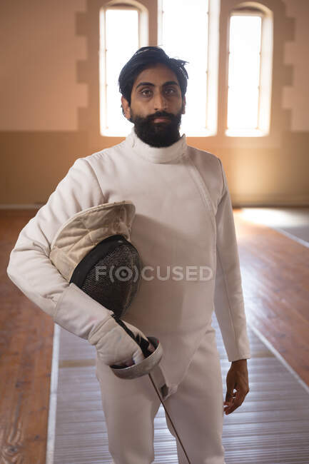 Portrait of mixed race sportsman wearing protective fencing outfit during a fencing training session, looking at camera, holding an epee. Fencers training at a gym. — Stock Photo