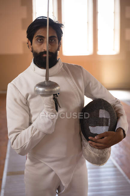 Portrait of mixed race sportsman wearing protective fencing outfit during a fencing training session, looking at camera, holding an epee. Fencers training at a gym. — Stock Photo