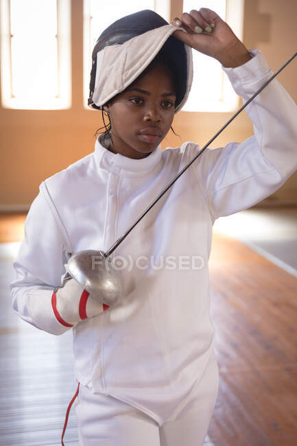 Femme sportive afro-américaine portant une tenue d'escrime protectrice lors d'une séance d'entraînement d'escrime, se préparant à un duel, tenant une épee et levant son masque. Entraînement des escrimeurs dans un gymnase. — Photo de stock