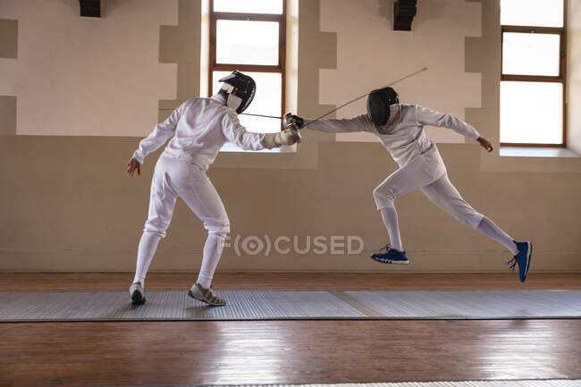 Kaukasische und gemischte Sportler in Schutzanzügen während eines Fechttrainings, zielen und stürzen sich mit ihren Degen aufeinander. Fechter beim Training im Fitnessstudio. — Stockfoto