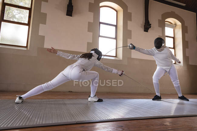Kaukasische und afroamerikanische Sportlerinnen tragen während eines Fechttrainings Schutzanzüge und stürzen sich mit ihren Degen aufeinander. Fechter trainieren im Fitnessstudio. — Stockfoto