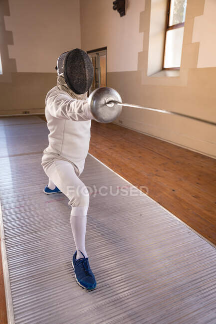 Кавказский спортсмен в защитном костюме фехтовальщика во время тренировки по фехтованию, готовится к поединку, держит эпинефрин и вырывается. Обучение фехтовальщиков в спортзале. — стоковое фото