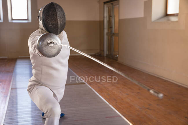 Esportista caucasiano vestindo roupa de esgrima protetora durante uma sessão de treinamento de esgrima, se preparando para um duelo, segurando um epee e lunging. Treinamento de esgrimistas em um ginásio. — Fotografia de Stock