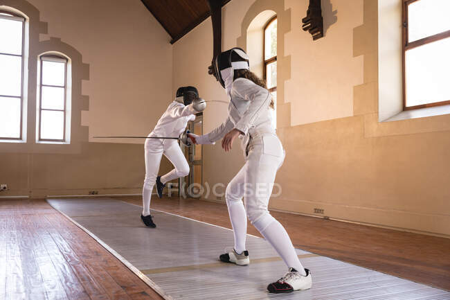 Caucasian and African American sportswomen wearing protective fencing outfit during a fencing training session, duelling with their epees. Fencers training at gym. — Stock Photo