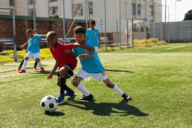 Вид спереди на смешанную расу и афроамериканского мальчика в командных костюмах, бегущего за мячом во время футбольного матча между двумя многонациональными командами мальчиков-футболистов на футбольном поле — стоковое фото