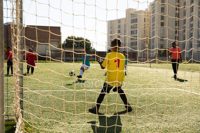 Vista trasera de un niño caucásico jugando en el gol con dos equipos multiétnicos de jugadores de fútbol de niño usando sus tiras de equipo, en acción durante un partido de fútbol en un campo de fútbol - foto de stock