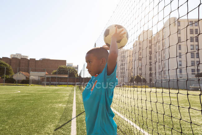 Vista lateral de un joven jugador de fútbol de raza mixta usando su tira de equipo, en acción durante un partido de fútbol, lanzando la pelota al campo de fútbol - foto de stock