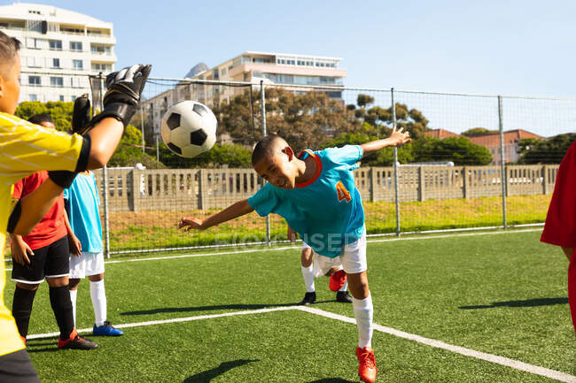 Передній вид змішаної раси, що очолює м'яч і Кавказького хлопчика, який грає з руками, щоб блокувати його, граючи з двома багатонаціональними командами футболістів, які грають у командні стрипи, під час футбольного матчу на футбольному полі. — стокове фото