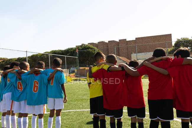 Вид сзади на две мультиэтнические команды мальчиков-футболистов, одетых в командные костюмы, стоящих на игровом поле, обнимающих друг друга — стоковое фото