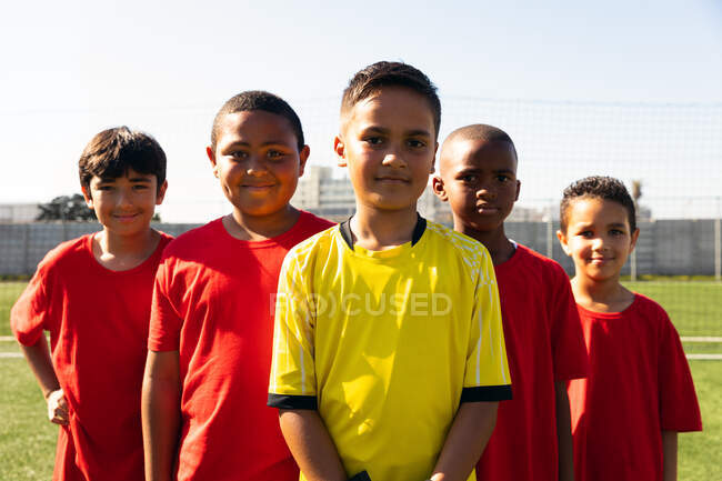 Вид спереду на групу молодих багатоетнічних футболістів-хлопчиків у командній смузі, що стоять на ігровому полі, посміхаючись — стокове фото