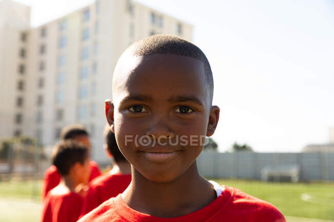Portrait of an African American boy soccer player wearing a team strip, standing on a playing field in the sub, looking to camera and smiling, with teammates standing in the background — Stock Photo