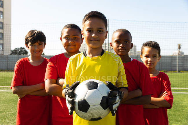 Front view of a multi-ethnic group of boy soccer players wearing their team strip, standing on a playing field on a sunny day with arms crossed, looking to camera and smiling, one of them holding the ball — Stock Photo