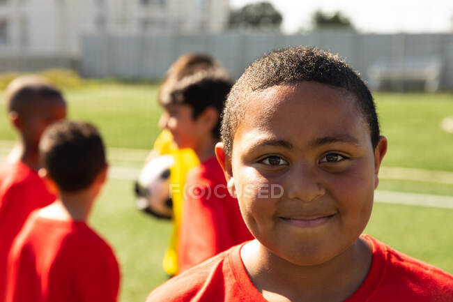Retrato de un futbolista de raza mixta usando su tira de equipo, parado en un campo de juego bajo el sol, mirando a la cámara y sonriendo, con compañeros de equipo de pie en el fondo - foto de stock
