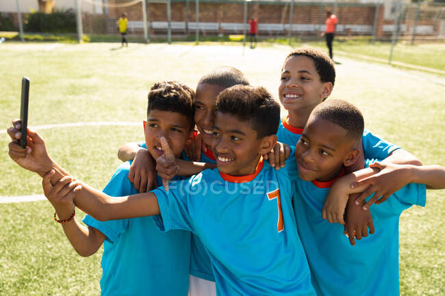 Vista lateral de un grupo multiétnico de jugadores de fútbol con su tira de equipo, de pie en un campo de juego tomando una selfie con un teléfono inteligente, posando con los brazos alrededor de los demás, sonriendo - foto de stock