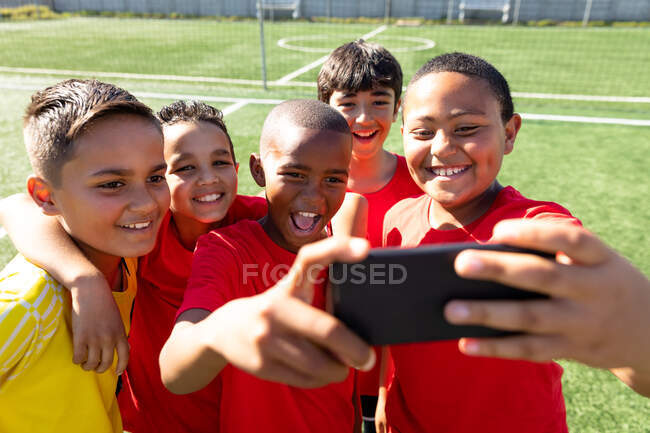 Vista frontale da vicino di un gruppo multietnico di giocatori di calcio maschili che indossano la loro striscia di squadra, in piedi su un campo da gioco facendo un selfie con uno smartphone, e posando con le braccia intorno a vicenda, sorridendo — Foto stock