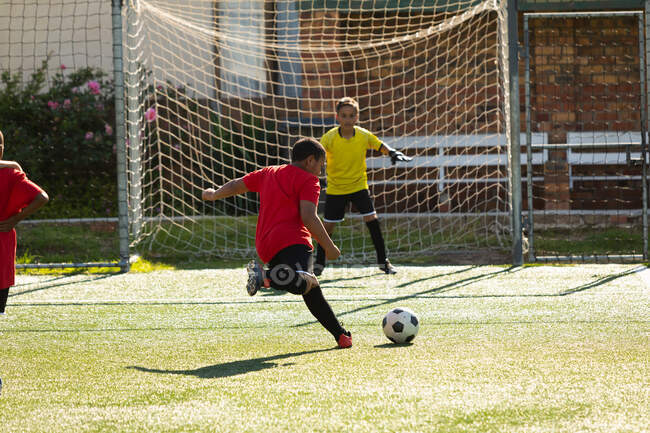 Rear view of a mixed race boy talking a shot at goal during a match between two teams of multi ethnic boy soccer players wearing their team strips on a playing field — Stock Photo