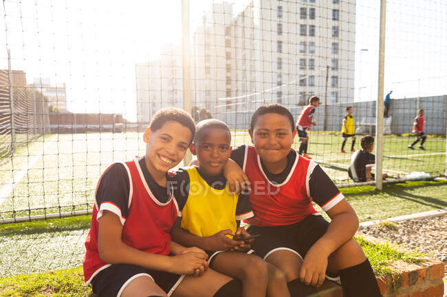 Retrato de un grupo multiétnico de tres jugadores de fútbol de niños que llevan su tira de equipo, sentados junto a un campo de juego, sosteniendo un teléfono inteligente, mirando a la cámara y sonriendo, con compañeros de equipo jugando fútbol en el fondo - foto de stock