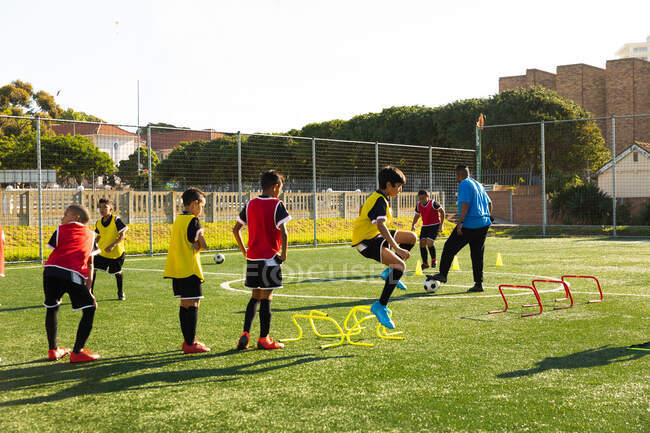 Вид сбоку на многонациональную группу мальчиков-футболистов, тренирующихся со своим тренером на игровом поле под солнцем — стоковое фото