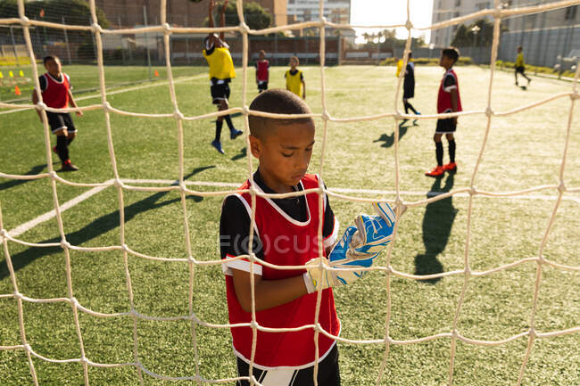 Front view of a young mixed race boy soccer player standing in goal and putting on goalkeepers gloves, preparing for a soccer match on a playing field in the sun, seen through the goal net, with teammates playing in the background — Stock Photo