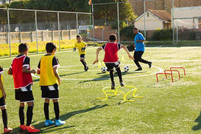 Rückansicht einer Gruppe multiethnischer Junge-Fußballer, die an einem sonnigen Tag mit ihrem Trainer auf einem Spielfeld trainieren und über niedrige Hürden springen — Stockfoto
