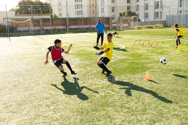 Vorderansicht einer Gruppe multiethnischer Junge-Fußballer, die an einem sonnigen Tag mit ihrem Trainer auf einem Spielfeld trainieren, Ballgeschicklichkeit üben und über niedrige Hürden springen — Stockfoto