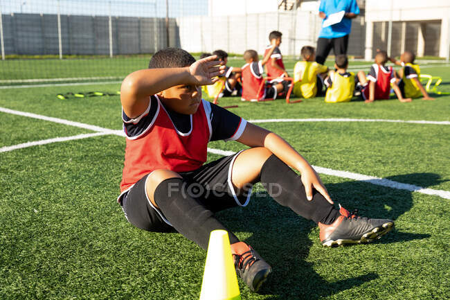 Вид збоку на змішану гонку хлопчик-футболіст сидить на ігровому полі на сонці, відпочиває під час тренування з футболу, одягаючи свою командну смугу, з товаришами по команді сидячи і слухаючи свого тренера на задньому плані — стокове фото