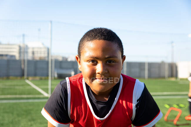 Портрет молодого змішаної гонки хлопчика-футболіста в червоній командній смузі, що стоїть на ігровому полі в сонячний день, дивлячись на камеру і посміхаючись — стокове фото