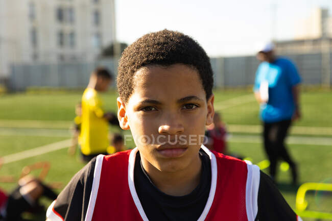 Porträt eines jungen Fußballspielers mit gemischter Rasse, der Mannschaftskleidung trägt, auf einem Spielfeld in der Sonne steht und in die Kamera blickt, im Hintergrund Teamkollegen und ihr Trainer — Stockfoto