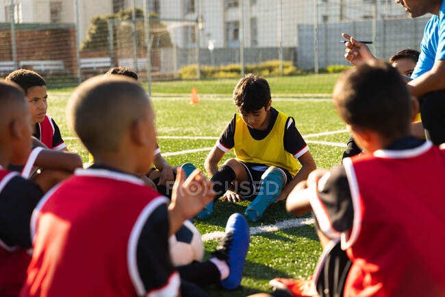 Vista lateral de un grupo multiétnico de jugadores de fútbol de niños sentados en un campo de juego en un día soleado escuchando a su entrenador masculino de raza mixta durante una sesión de entrenamiento de fútbol - foto de stock