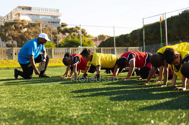 Side view of a mixed race male soccer coach kneeling and instructing a multi-ethnic group of boy soccer players doing press ups in a row on a playing field in the sun during a soccer training session — Stock Photo