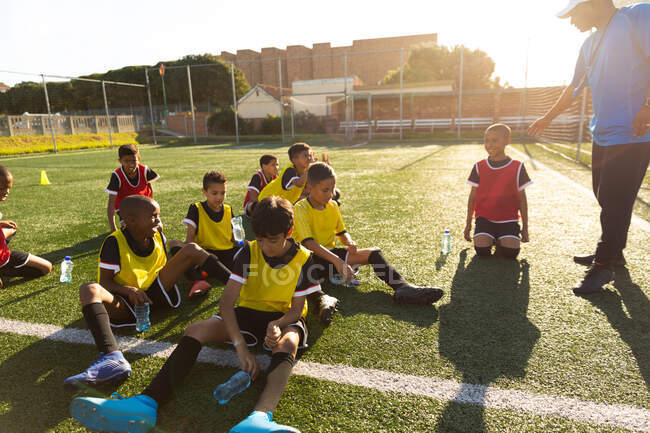 Side view of a mixed race male soccer coach standing and instructing a multi-ethnic group of boy soccer players sitting on a playing field in the sun during a soccer training session, backlit — Stock Photo