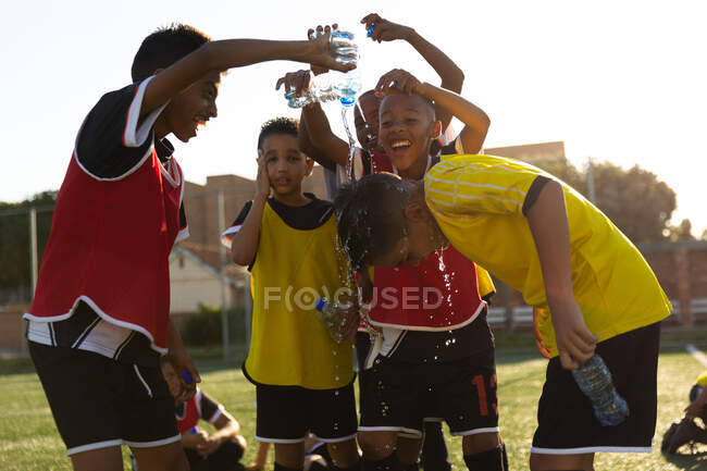 Seitenansicht einer multiethnischen Gruppe von Jungen-Fußballern auf einem Spielfeld in der Sonne, die sich abkühlen und Spaß haben, sich gegenseitig Wasser aus Flaschen übergießen und beim Fußballtraining lachen. — Stockfoto