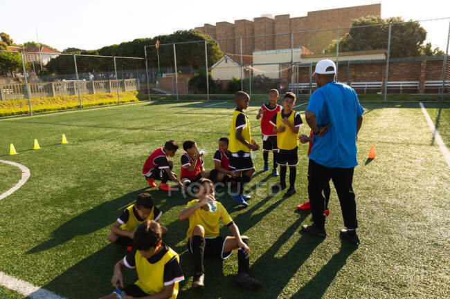 Вид спереду змішаної раси чоловічий тренер з футболу стоїть і навчає багатоетнічній групі хлопчиків футболістів, які сидять на ігровому полі на сонці під час тренування футболу — стокове фото
