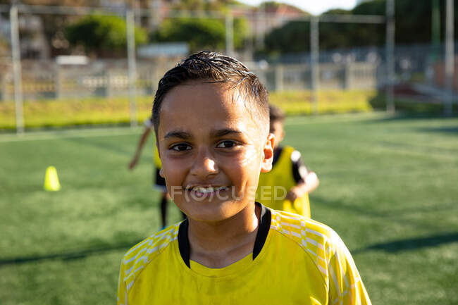 Porträt eines jungen Fußballspielers mit gemischter Rasse, der einen gelben Mannschaftsstreifen trägt, in die Kamera blickt und lächelt, auf einem Spielfeld in der Sonne steht, mit Teamkollegen im Hintergrund — Stockfoto