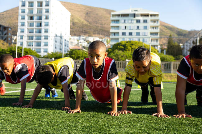 Vista frontal de un grupo multiétnico de jugadores de fútbol de niños haciendo flexiones de presión en una fila en un campo de juego al sol durante una sesión de entrenamiento de fútbol - foto de stock