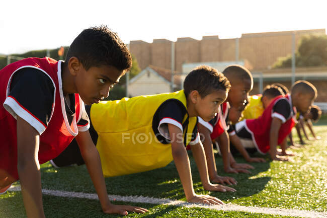 Vista laterale da vicino di un gruppo multietnico di giocatori di calcio maschili che fanno press up di fila su un campo da gioco al sole durante una sessione di allenamento di calcio — Foto stock