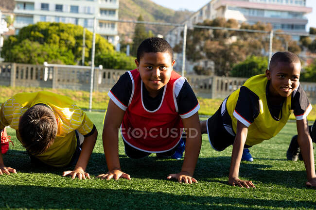 Front view close up of a multi-ethnic group of boy soccer players doing press ups in a row on a playing field in the sun during a soccer training session — Stock Photo