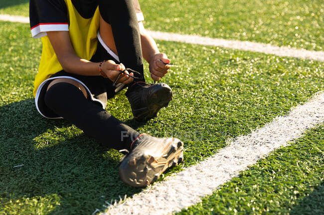 Vista frontal de la sección baja del jugador de fútbol niño sentado en un campo de fútbol al sol poniéndose sus botas de fútbol y atando los cordones durante una sesión de entrenamiento - foto de stock