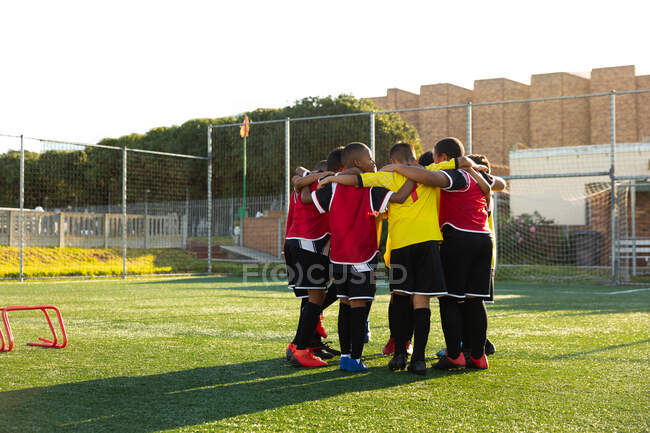 Side view of a multi-ethnic group of boy soccer players standing in a huddle on a playing field in the sun, embracing each other and celebrating winning the match — Stock Photo