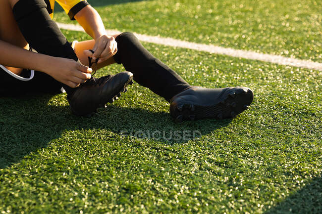Side view low section of boy soccer player sitting on a soccer pitch in the sun putting on his football boots and tying the laces up during a training session — Stock Photo