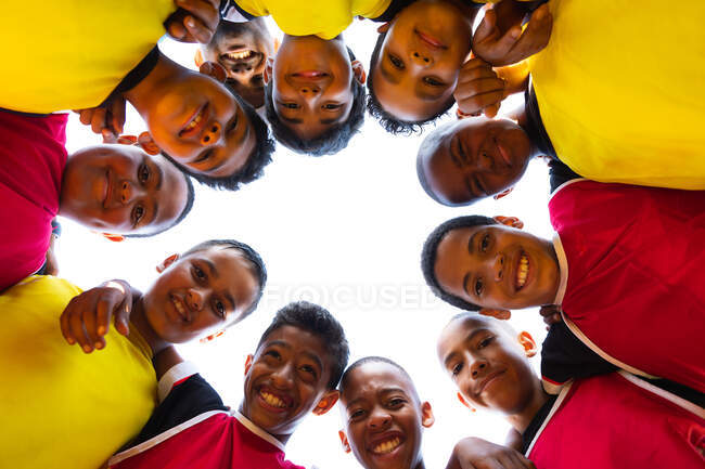 Низкий угол приближения многонациональной группы мальчиков-футболистов, стоящих в мотивационной толпе на игровом поле на солнце, обнимающих друг друга, обнимающих друг друга и смотрящих вниз на камеру улыбающихся и смеющихся перед матчем — стоковое фото