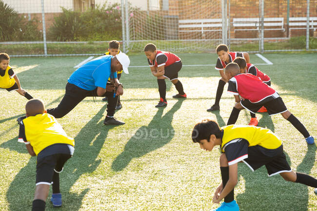 Вид сбоку на тренера смешанной расы мужского пола, стоящего и инструктирующего многонациональную группу мальчиков-футболистов, делающих упражнения на игровом поле на солнце во время тренировки по футболу — стоковое фото