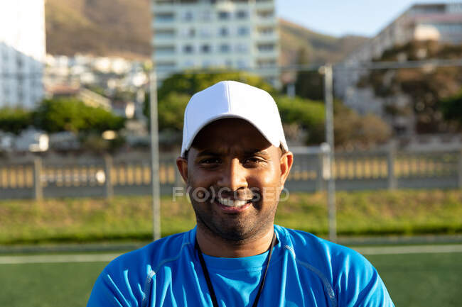 Retrato de cerca de un entrenador de fútbol masculino de raza mixta con una gorra de béisbol, de pie en un campo de juego al sol, mirando a la cámara y sonriendo durante una sesión de entrenamiento de fútbol - foto de stock