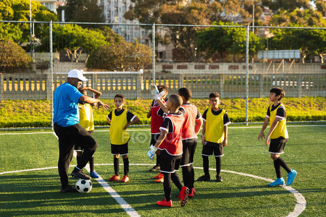 Seitenansicht eines gemischten männlichen Fußballtrainers, der mit dem Fuß auf einem Fußball steht und während eines Fußballtrainings eine multiethnische Gruppe von Jungen-Fußballern auf einem Fußballplatz in der Sonne zeigt und unterrichtet. — Stockfoto